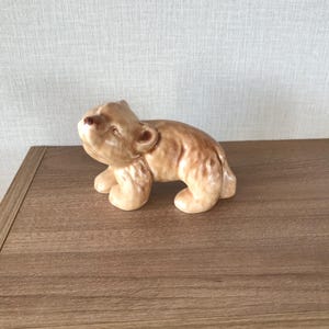 May include: A ceramic bear figurine in shades of tan and brown. The bear is posed on all fours, with its head tilted upwards. The figurine has a glossy finish and is set on a wooden surface.
