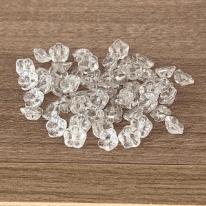May include: A collection of small, clear, flower-shaped beads. Each bead features a five-petalled design with a central hole, crafted from a transparent material that reflects light, creating a sparkling appearance across the group of loose items.