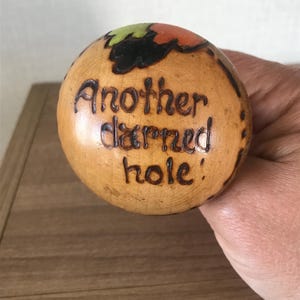 May include: A wooden ball with a burnt-in design of leaves and the words "Another darned hole!"
