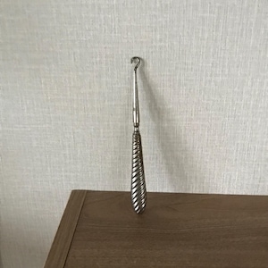 May include: A silver crochet hook with a twisted handle. The hook is placed on a brown wooden surface against a textured white wall. The hook's design is simple and elegant, suitable for various crafts.