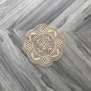 May include: A beige, circular doily with an intricate lace design. The doily features a central floral pattern surrounded by detailed openwork and scalloped edges. The delicate textile is placed on a gray wooden surface.