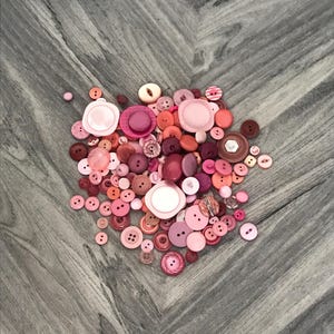May include: A collection of various pink buttons in different shades and sizes. The buttons are scattered on a gray wooden surface, showcasing a range of shapes, including round, flat, and domed designs. Some buttons have two or four holes, while others have a shank.