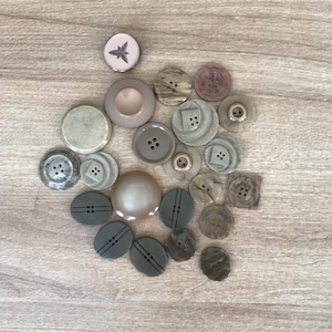 May include: A collection of various buttons in shades of gray, beige, and brown, arranged on a light wood surface. The buttons vary in size, shape, and design, including round, oval, and square options. Some have multiple holes, while others feature decorative details.