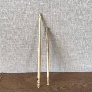 May include: Two light-colored wooden knitting needles. One needle is longer than the other. Both needles have a series of carved rings near the bottom. The needles are set against a light gray textured background and a brown wooden surface.