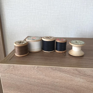 May include: Five wooden spools of thread, each with a different color of thread. The spools are arranged in a row on a wooden surface. The colors of the thread are brown, white, black, red, and white.