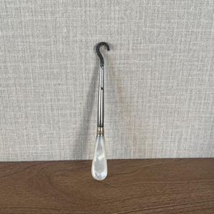 May include: A button hook featuring a curved metal tip and a tapered, iridescent white mother-of-pearl handle. The tool has a slender, polished metal shaft connecting the hook to the smooth, teardrop-shaped handle, which displays a natural luster.