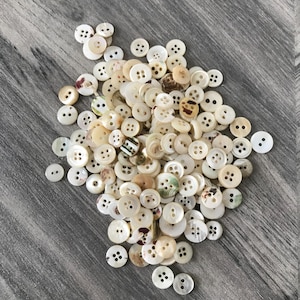 Peut inclure: Un ensemble de boutons blanc cassé et crème à quatre trous, dispersés sur une surface en bois grise. Certains boutons ont un éclat nacré, tandis que d'autres présentent de subtiles variations de couleur. Les boutons varient en taille et en forme, pour la couture et les loisirs créatifs.