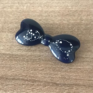May include: A navy blue bow-shaped hair clip with white dot detailing. The glossy accessory is set against a wooden surface. The bow has a rounded shape and a smooth, reflective surface, adding a touch of elegance.