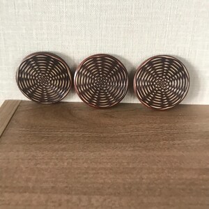 May include: Three round brown and white coasters with a woven pattern. The coasters are arranged in a row on a wooden surface.