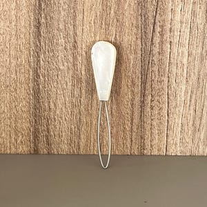 May include: A button hook tool featuring a smooth, pearlescent white, teardrop-shaped handle attached to a thin, flexible, silver-toned metal wire loop. The handle has a glossy finish with subtle marbled texture, while the wire loop is elongated.