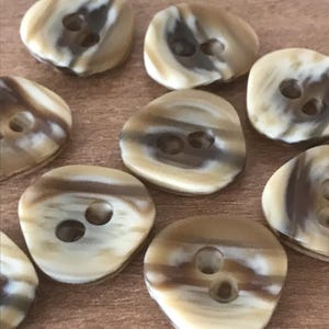 May include: A collection of oval-shaped buttons with two holes each. The buttons have a marbled appearance with shades of tan, cream, and brown. The buttons are scattered across a wooden surface.