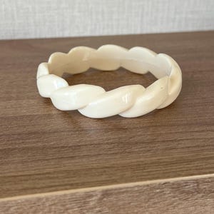 May include: A solid off-white bangle bracelet featuring a continuous twisted rope design. The circular accessory has a smooth, glossy finish and a uniform thickness throughout its entire structure, creating a simple and cohesive appearance.