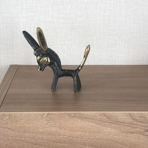 May include: A small, bronze donkey figurine with gold-colored accents on the ears and tail. The donkey is posed in a standing position, with its head slightly lowered. The figurine is placed on a wooden surface.