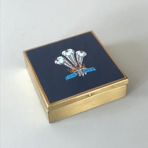 May include: A small, square, gold-colored box with a blue lid. The lid has a white image of three feathers with a crown and a blue ribbon with the words "ICH DIEN" on it.