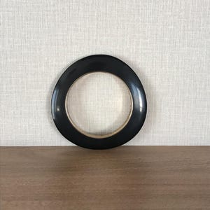 May include: A black, circular object with a gold-coloured inner ring. The object is centred on a wooden surface, with a textured, light-coloured wall in the background. The object's shape is a thick ring.