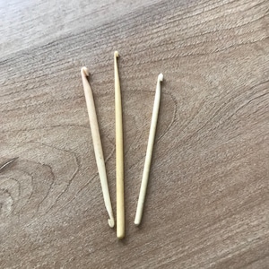 May include: Three wooden crochet hooks of varying sizes and shades of cream and beige are arranged on a wooden surface. The hooks are slender, with a curved end for catching yarn. The wooden surface has a natural wood grain pattern.