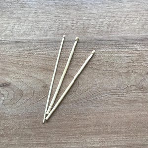 May include: Three off-white crochet hooks of varying lengths are arranged on a light brown wooden surface. The hooks are slender, with a hook at one end and a slightly thicker handle on one of them. The wooden surface has a natural grain pattern.
