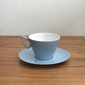 May include: A pale blue teacup and saucer set. The teacup has a curved shape with a handle and a white interior. The saucer is a matching pale blue. The set is on a wooden surface.