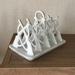 May include: A white ceramic toast rack with six slots, each featuring a decorative design. The rack sits on a rectangular base with gold accents. The design is a series of arches and curves.