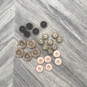May include: Assortment of buttons in various shades of brown and beige. The buttons are round and have two holes. There are 15 buttons in total.