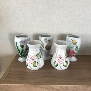 May include: Five white ceramic vases with floral designs. The vases are arranged on a wooden surface. The designs include a thistle, a cluster of pink flowers, and a red flower with green leaves.