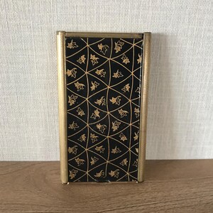 May include: A small, rectangular, black and gold metal box with a decorative pattern of gold leaves on a black background. The box has a gold frame around the edges.
