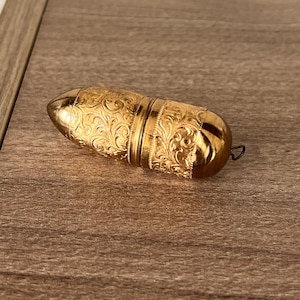 May include: A gold-toned metal pill case with a cylindrical, bullet-like shape. The surface features an intricate, embossed scrollwork pattern throughout the body. It has a smooth, pointed end and a small metal loop attached to the rounded base.