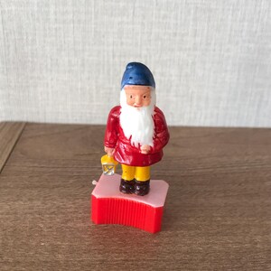 May include: A red and white plastic pencil sharpener shaped like a dwarf. The dwarf is wearing a red jacket, yellow pants, and a blue hat. He has a white beard and is holding a lantern.