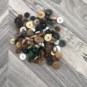 May include: A large collection of assorted buttons in various colors, including black, white, brown, beige, and green. The buttons are made of different materials, such as plastic, metal, and wood. Some buttons have two holes, while others have four holes.