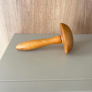May include: A vintage wooden darning mushroom with a smooth, light brown finish. The tool consists of a rounded, dome-shaped top attached to a tapered, cylindrical handle, showing natural wood grain patterns and signs of light wear.