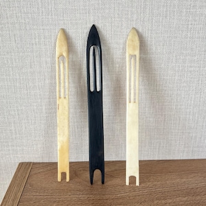 May include: Three net needles arranged vertically, featuring two off-white needles on the sides and one black needle in the centre. Each needle has a pointed tip, a long central slot, and a U-shaped notch at the base for holding fishing line.