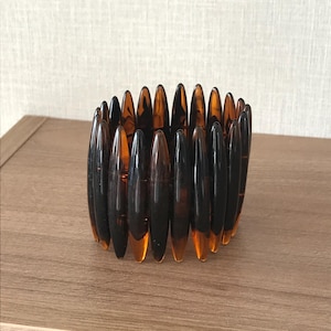 May include: A brown and amber stretch bracelet. The bracelet features elongated, pointed beads. The beads are arranged in a circular pattern, creating a bold accessory. The bracelet is displayed on a wooden surface.