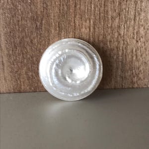 May include: A clear, circular glass object with concentric rings, likely a lid or stopper. The object is centered against a brown wooden background and a gray surface.