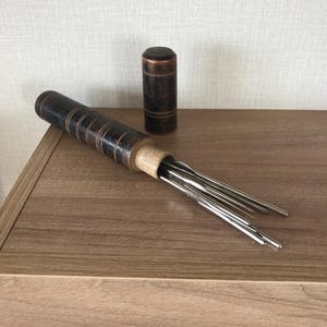 May include: A wooden needle case with a dark brown finish and metal needles. The case is cylindrical with decorative rings. A smaller, matching wooden cylinder stands nearby. The items are on a wooden surface.