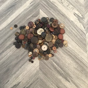 May include: A large collection of brown, beige, and black buttons of various sizes and shapes. The buttons are scattered on a light gray wood surface.