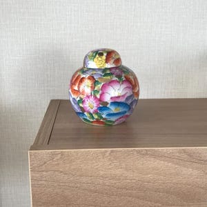 May include: A small, round ceramic jar with a lid, adorned with a vibrant floral pattern in shades of pink, orange, blue, and yellow. The jar sits on a wooden surface, showcasing a decorative piece.