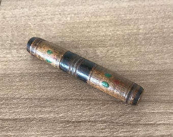 May include: A wooden pen holder with a brown finish, accented with black and green details. The pen holder is cylindrical and appears to be handmade. The pen holder is lying on a wooden surface.
