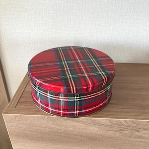 Könnte beinhalten: Eine runde, rote Dose mit einem Schottenmuster in Grün, Gelb, Blau und Weiß. Die Dose steht auf einem Holzregal. Deckel und Seiten der Dose weisen das gleiche Karomuster auf.