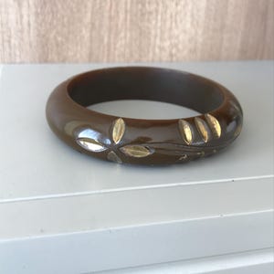 May include: A brown, round bangle bracelet with a floral design. The bracelet is a dark brown color with gold-colored leaf accents. The bracelet is made of a solid material and has a smooth, polished finish.