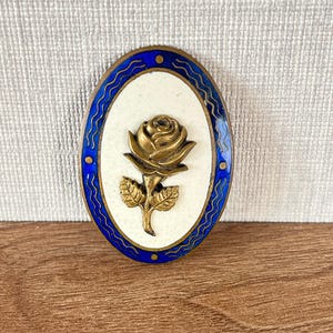 May include: An oval brooch featuring a central brass rose relief set against an off-white enamel background. The border is composed of deep blue enamel with a wavy gold-toned line pattern and four small circular brass accents at the cardinal points.