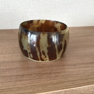 May include: A wide, round bracelet with a marbled pattern of brown and tan. The bracelet is made of a glossy material and has a smooth, polished finish. The design resembles natural stone.