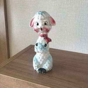 May include: A vintage ceramic lamb figurine with a whimsical design. The lamb has a white body with blue accents, pink ears, and a red scarf. The figurine has black eyes and feet, and is likely a collectible or decorative item.