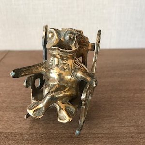 May include: A brass frog ornament sitting in a rocking chair. The frog has detailed features and is wearing a collar. The rocking chair has a classic design. The ornament is a decorative item.