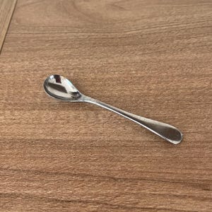 May include: A small, polished silver-coloured metal spoon with a rounded bowl and a smooth, tapered handle. The spoon features a reflective surface and a simple, clean design, showcasing a classic utensil shape with no additional patterns or markings.