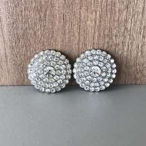 May include: Two silver-toned, round buttons with a floral design. Each button features a large, clear rhinestone in the center, surrounded by smaller, sparkling rhinestones. The buttons are set against a wooden background.