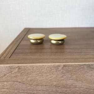 May include: Two small, round, white and gold drawer pulls with a smooth, shiny finish. The pulls are on a wooden surface.