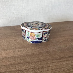May include: A small, round, white porcelain trinket box with a floral design. The lid has a scalloped edge and features a blue and white floral pattern. The box is decorated with a variety of flowers, including yellow, red, and blue.
