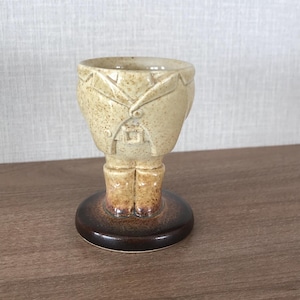 May include: A ceramic egg cup in the shape of a person, with a tan jacket and brown boots. The egg cup has a round, dark brown base. The design includes details like a lapel, buttons, and a belt buckle.