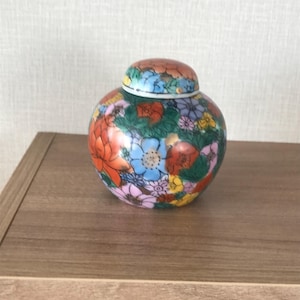 May include: A round, ceramic jar with a lid, adorned with a vibrant floral pattern. The jar features a base color of dark green, with colorful flowers in shades of red, orange, blue, pink, and yellow. The lid mirrors the floral design.
