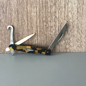 May include: A small, folding pocket knife with a tortoiseshell-patterned handle. The knife has a blade, a hook, and a small, pointed tool.